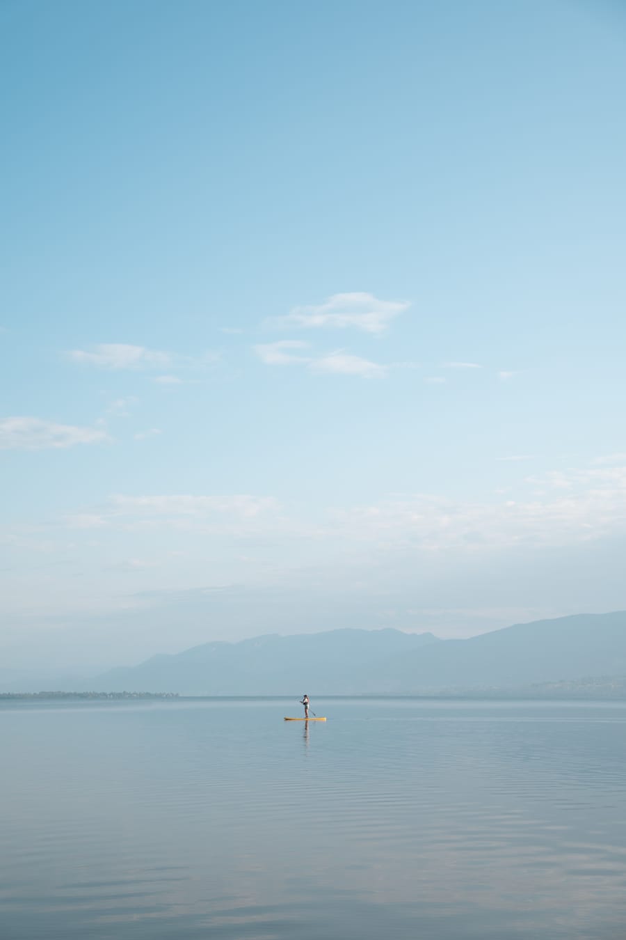 A lone paddleboarder on calm water with hills in the haze